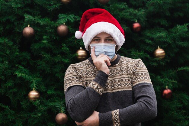 Idea natalizia. Close up Ritratto di uomo che indossa un cappello di Babbo Natale, maglione di Natale e mascherina medica con emozione. Sullo sfondo di un albero di Natale. Pandemia di coronavirus