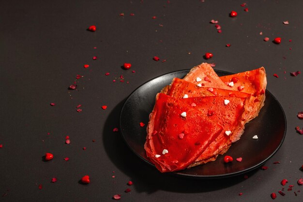 Idea romantica per la colazione, crepes rosse o frittelle sottili ricoperte di cuori dolci. Cibo festivo per San Valentino o festa della mamma. Luce dura alla moda, ombra scura. Sfondo cemento pietra nera, copia spazio