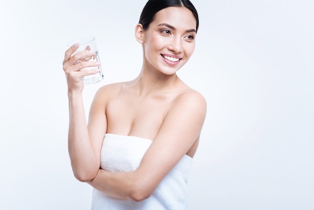 Idratazione cruciale. Bella giovane donna ottimista in un asciugamano in posa con un bicchiere d'acqua e sorridente, guardando in lontananza, mentre in piedi isolato su un muro bianco