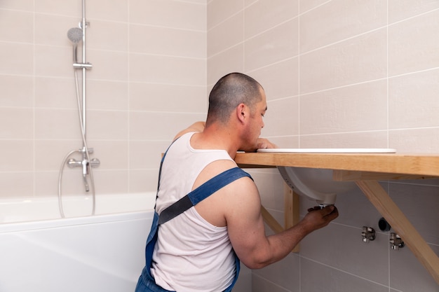 Idraulico in tuta di jeans blu che ripara lavandino in bagno con parete di piastrelle