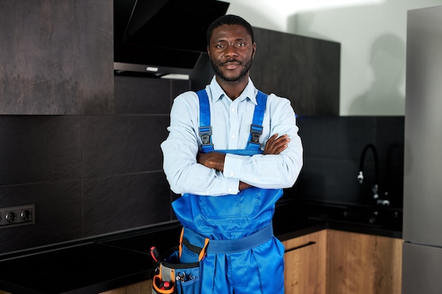 Idraulico tuttofare africano bello serio in uniforme blu. Giovane abile tuttofare in generale in piedi a casa in cucina in posa guardando la fotocamera. Servizio di riparazione, concetto di duro lavoro