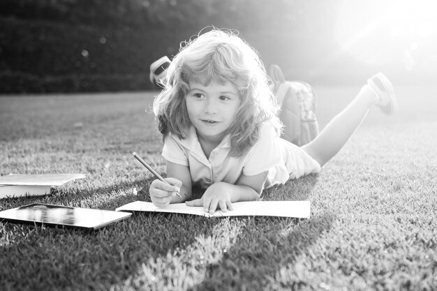 Il bambino adorabile sveglio con la scrittura della matita sul taccuino fuori il libro legge il libro del bambino nel parco