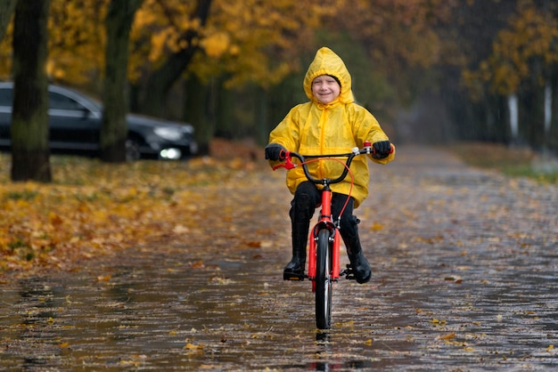 Il bambino allegro in impermeabile giallo va in bicicletta nel parco autunnale. Il ragazzo sta guidando lungo un vicolo in un parco piovoso.