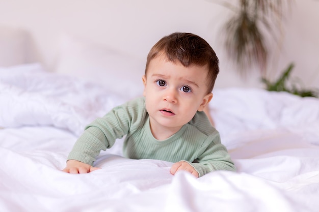 Il bambino allegro in un body di cotone verde è sdraiato a pancia in giù su un letto bianco