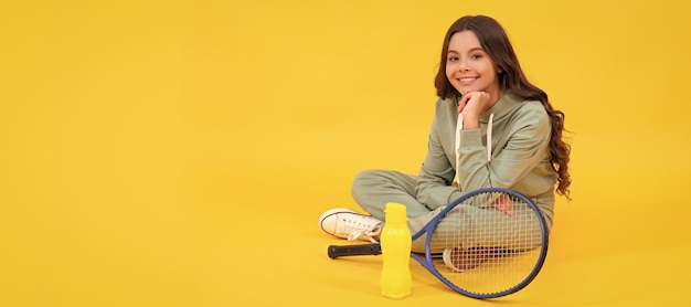 Il bambino allegro si siede in abbigliamento sportivo con la racchetta da badminton e la bottiglia d'acqua su sfondo giallo si rilassano