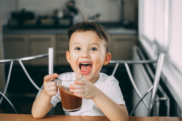 il bambino beve cioccolato che si dimena in cucina durante il giorno, tiene una tazza tra le mani e urla