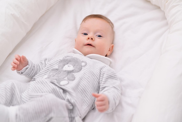 Il bambino biondo ridente giace in una culla sulla schiena in una stanza a casa con lenzuola di cotone bianco