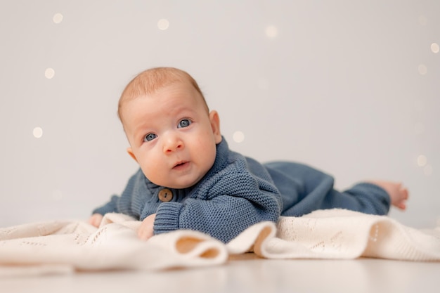 Il bambino carino con gli occhi azzurri in tuta lavorata a maglia blu giace sulla pancia su uno sfondo solido scozzese grigio