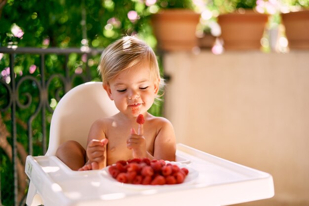 Il bambino carino si siede su un seggiolone mettendo i lamponi al dito