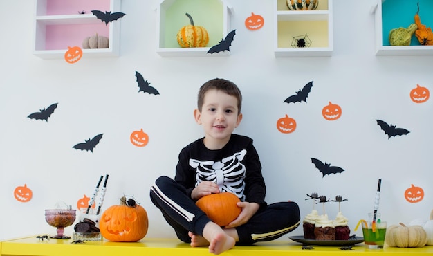 Il bambino caucasico in costume da scheletro tiene la zucca ricurva e si siede sul tavolo con cibo e decorazioni di Halloween