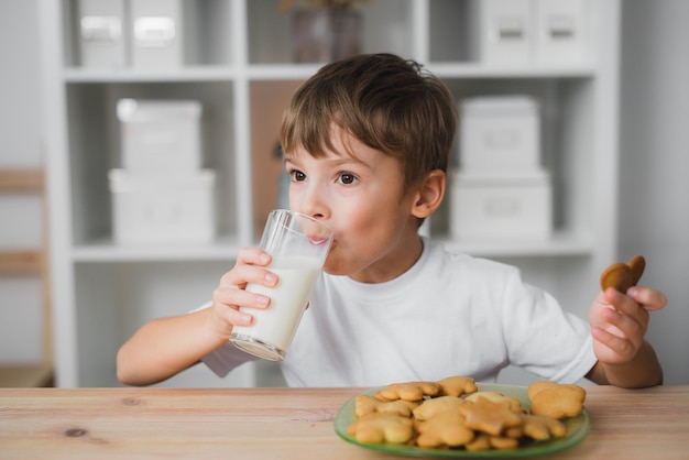 Il bambino caucasico si siede al tavolo, beve latte e mangia i biscotti.