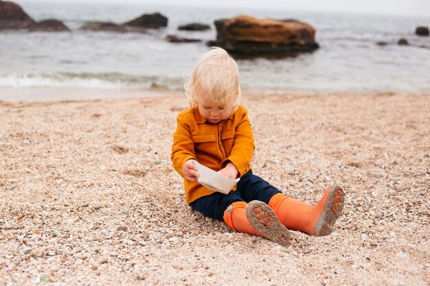 Il bambino che gioca con la barchetta di carta in spiaggia in autunno