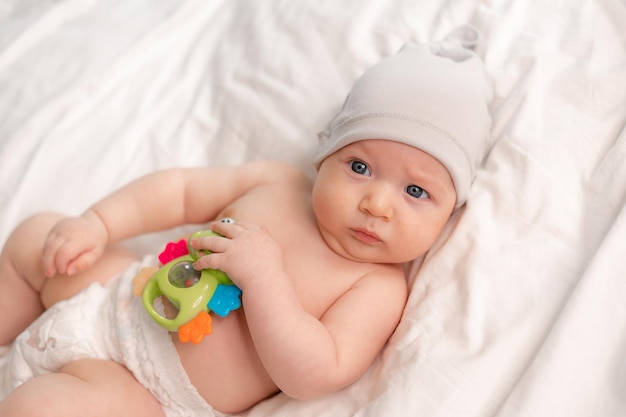 Il bambino con un pannolino e un berretto bianco gioca presto con un giocattolo verde sdraiato sulla schiena su lenzuola bianche