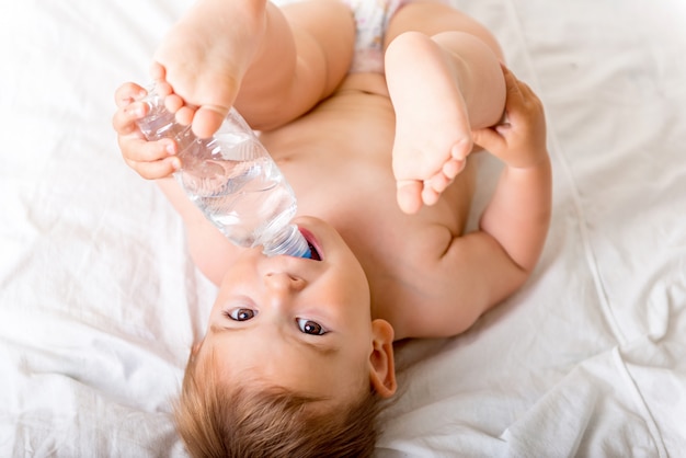 Il bambino del bambino che pone sul letto bianco, sorride e beve l&#39;acqua dalla bottiglia di plastica