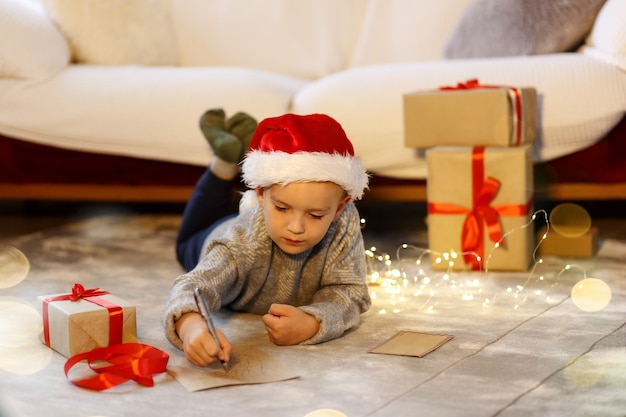 Il bambino di Natale scrive una lettera a Babbo Natale con il cappello di Natale scrive la lista dei desideri accanto all'albero di Natale