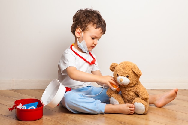 Il bambino divertente in una mascherina medica gioca con un orsacchiotto dal medico.