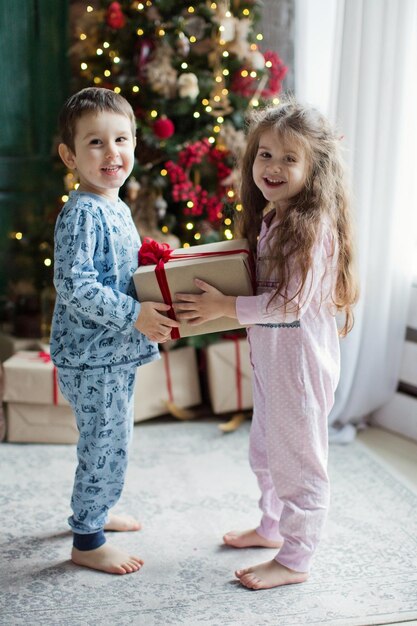 Il bambino e la ragazza in pigiama tengono scatole regalo vicino all'albero di Natale alla luce Interni natalizi Bambini di Natale carini