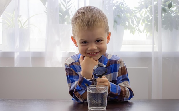 Il bambino esamina l'acqua con una lente d'ingrandimento in un vetro Kid