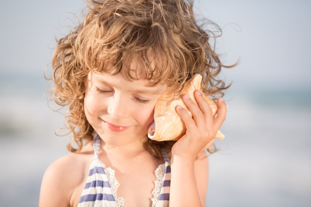 Il bambino felice ascolta la conchiglia in spiaggia Concetto di vacanze estive