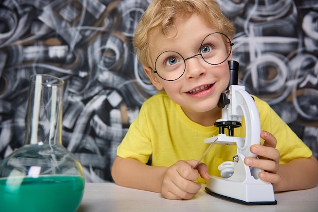 Il bambino felice con gli occhiali con enormi occhi azzurri usa un vero microscopio