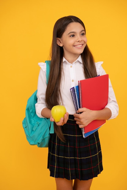 Il bambino felice con lo zaino e le cartelle di lavoro tiene il pranzo alla mela in uniforme scolastica su sfondo giallo, settembre.