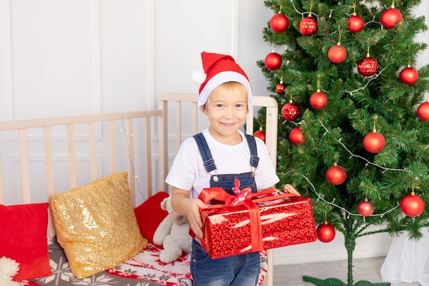 Il bambino felice con un cappello rosso tiene un regalo vicino all'albero di Natale