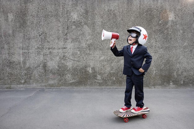 Il bambino felice finge di essere un uomo d'affari. Bambino divertente che guida lo skateboard all'aperto. Ragazzo che grida attraverso l'altoparlante. Ritorno al lavoro, avvio e concetto di idea imprenditoriale