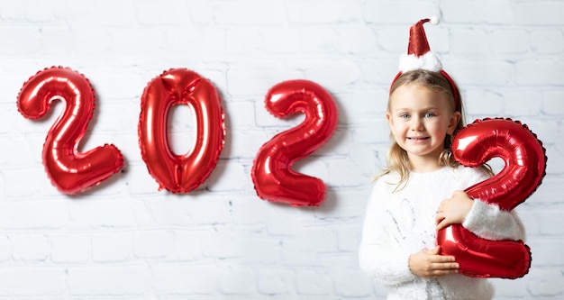 Il bambino felice in cappello di Babbo Natale ha isolato il fondo bianco della parete della rottura figure palloncini i numeri del nuovo anno 2022