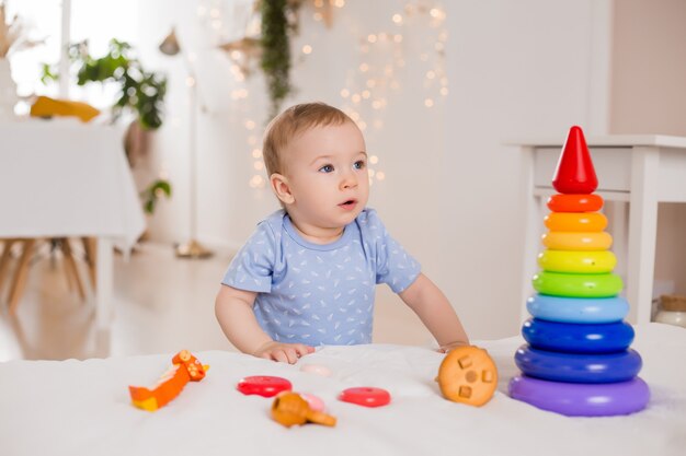 Il bambino felice in tuta blu si siede a casa sul letto che gioca con i giocattoli di sviluppo