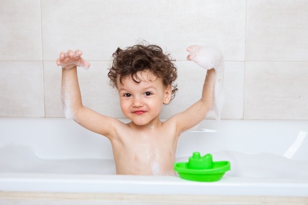 Il bambino felice si bagna in un bagno mostra la schiuma nelle sue mani