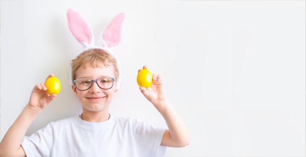 Il bambino felice sorride con gli occhiali in una maglietta bianca con le orecchie di coniglio e con le uova di Pasqua gialle colorate