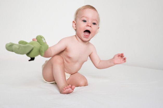 Il bambino felice sta giocando con il peluche verde sul letto bianco nella stanza bianca