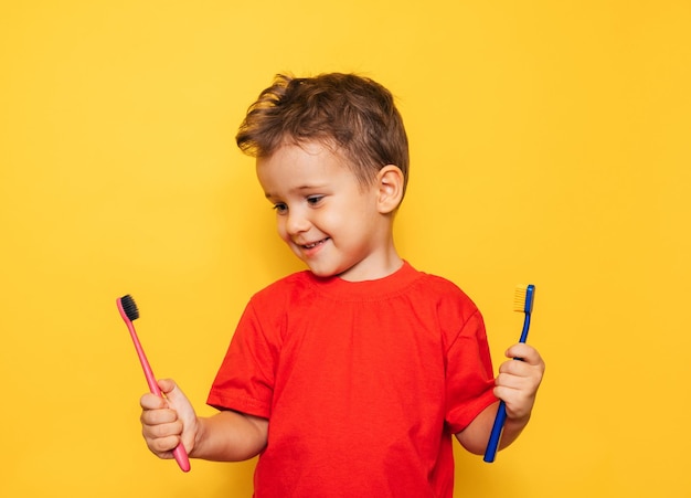 Il bambino felice tiene 2 spazzolini da denti su uno sfondo giallo con entrambe le mani e sorride mostrando i denti. Assistenza sanitaria, igiene orale.