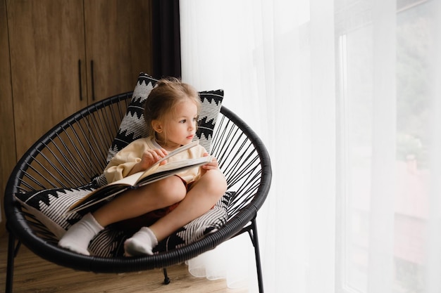 Il bambino guarda le pagine di un libro seduto su una sedia sul balcone dell'appartamento durante il giorno