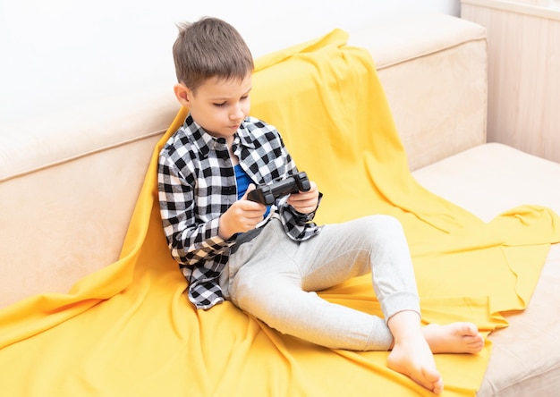 Il bambino in camicia a quadri seduto sul divano con il joystick nero nelle sue mani giocando al videogioco Giocare ai videogiochi a casa concetto