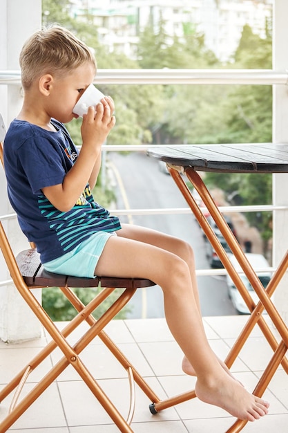 Il bambino in età prescolare beve latte caldo dalla tazza seduto sulla sedia al mattino presto sul balcone dell'hotel