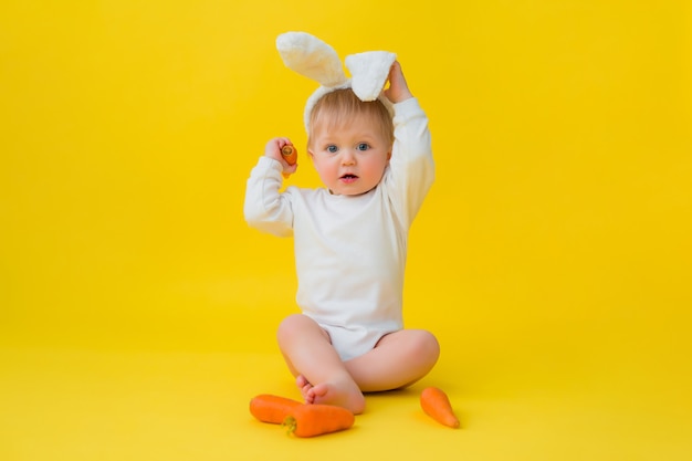 Il bambino in un body bianco con orecchie di coniglio in testa mangia carote, si siede su uno sfondo giallo con verdure. bambino sotto forma di un coniglietto di Pasqua, spazio per il testo