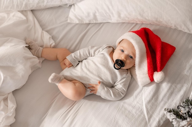 Il bambino in un body bianco e un cappello da Babbo Natale con il ciuccio nero in bocca è sdraiato nella vista dall'alto del letto