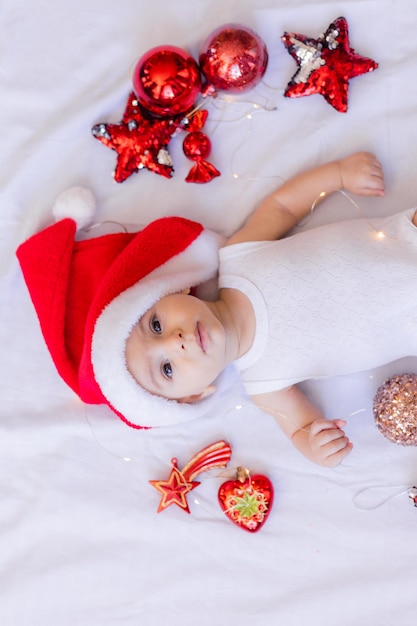 Il bambino in un body bianco e un cappello da Babbo Natale è sdraiato sulla schiena su un lenzuolo bianco circondato da giocattoli rossi dell'albero di Natale. inverno, capodanno. spazio per il testo. Foto di alta qualità