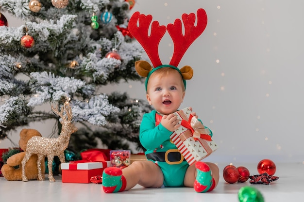 Il bambino in un body verde con le corna di cervo in testa sta giocando con scatole regalo vicino all'albero di Natale. bambino con un costume da gnomo di Natale. concetto di Capodanno invernale. merci per le vacanze. spazio per il testo