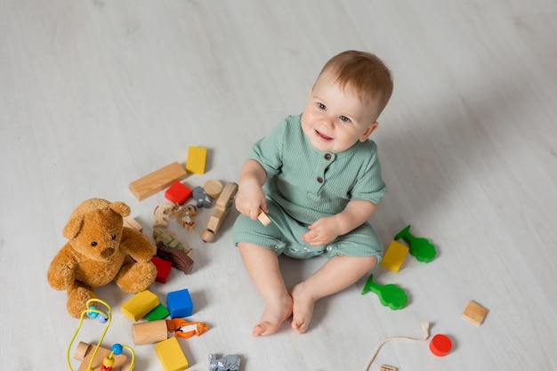 Il bambino in un body verde è seduto sul pavimento di legno della casa a giocare a giocattoli di legno educativi