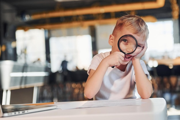 Il bambino intelligente in abiti casual con il laptop sul tavolo si diverte con la lente d'ingrandimento