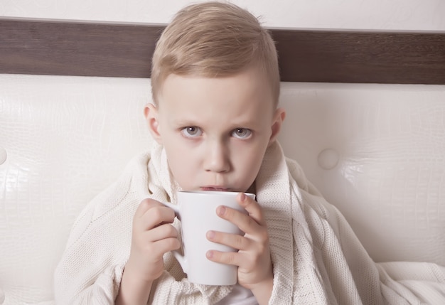 Il bambino malato seduto in un letto e tiene una tazza di tè. Concetto di medicina