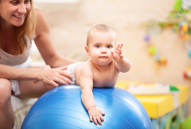 Il bambino nel pannolino sta rotolando su una palla da ginnastica blu con l'aiuto della mamma.