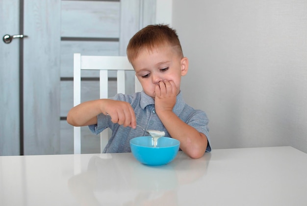 Il bambino non vuole mangiare e spinge via il piatto, un bambino è in cucina. Un bambino triste non vuole mangiare il porridge. Un bambino mangia senza appetito. TConcept di sana colazione per bambini.