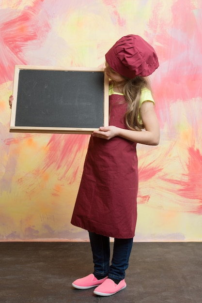 Il bambino o la bambina felice cucinano in cappello da cuoco rosso e grembiule tengono la lavagna del menu su sfondo colorato cucinando e mangiando lo spazio della copia