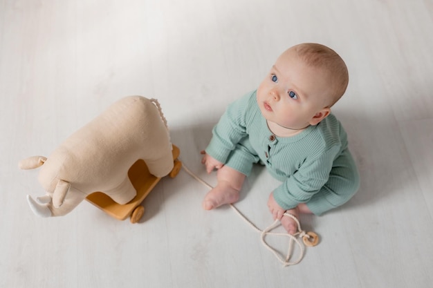 il bambino paffuto in una tuta verde è seduto sul pavimento e gioca con un giocattolo di legno su una corda