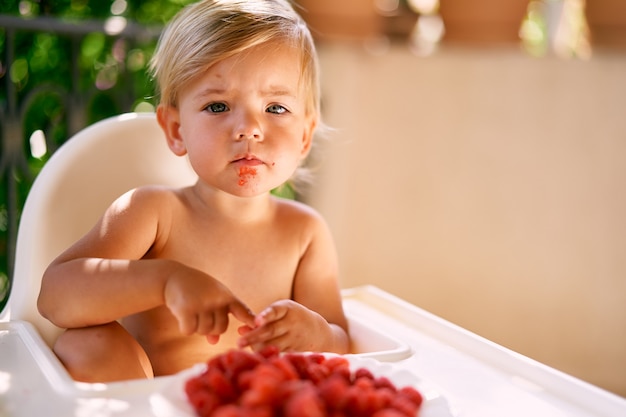 Il bambino pensieroso si siede su un seggiolone davanti a una ciotola di lamponi