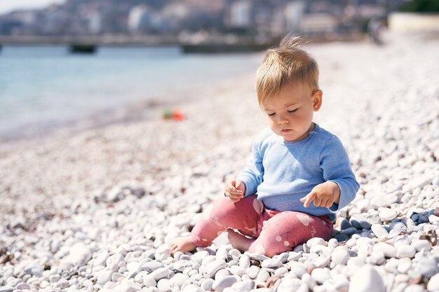 Il bambino pensieroso si siede su una spiaggia di ciottoli guardando i suoi piedi vicino al mare