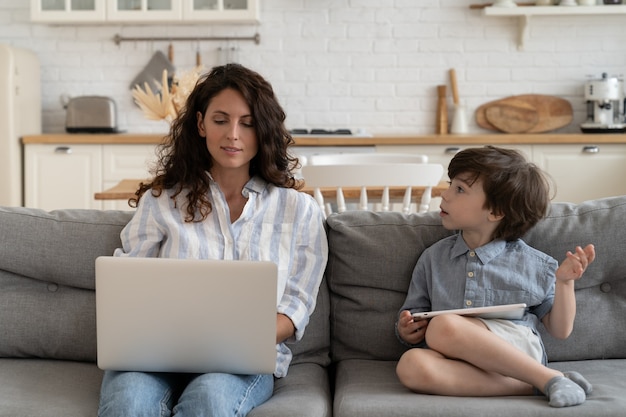 Il bambino piccolo parla con la mamma impegnata a digitare sul laptop mamma multitasking imprenditrice lavora da casa con figlio